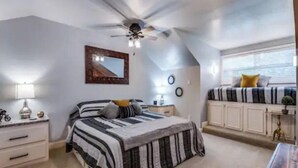 Un dormitorio con cama, ventilador de techo, mesita de noche, cómoda, ventana con vistas y una pequeña mesa con lámpara.