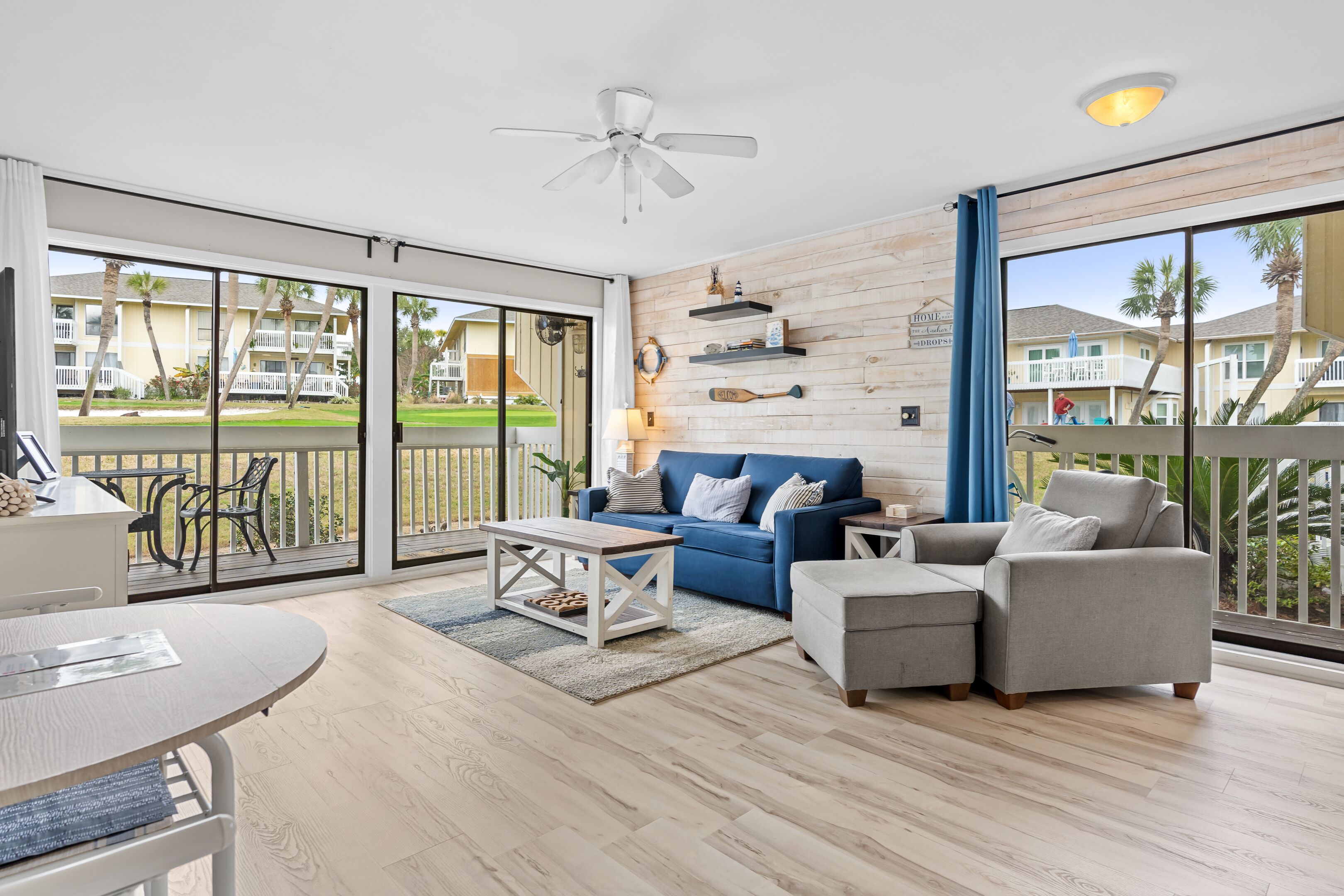A spacious living room with a blue sofa, grey armchair, and a white coffee table. There is a ceiling fan, wooden wall decor, and a view of a pool and buildings through large sliding glass doors.