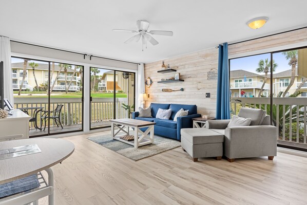 A spacious living room with a blue sofa, grey armchair, and a white coffee table. There is a ceiling fan, wooden wall decor, and a view of a pool and buildings through large sliding glass doors.