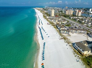 An aerial view of a beach with rows of umbrellas, beachfront buildings, and a clear view of the ocean.
