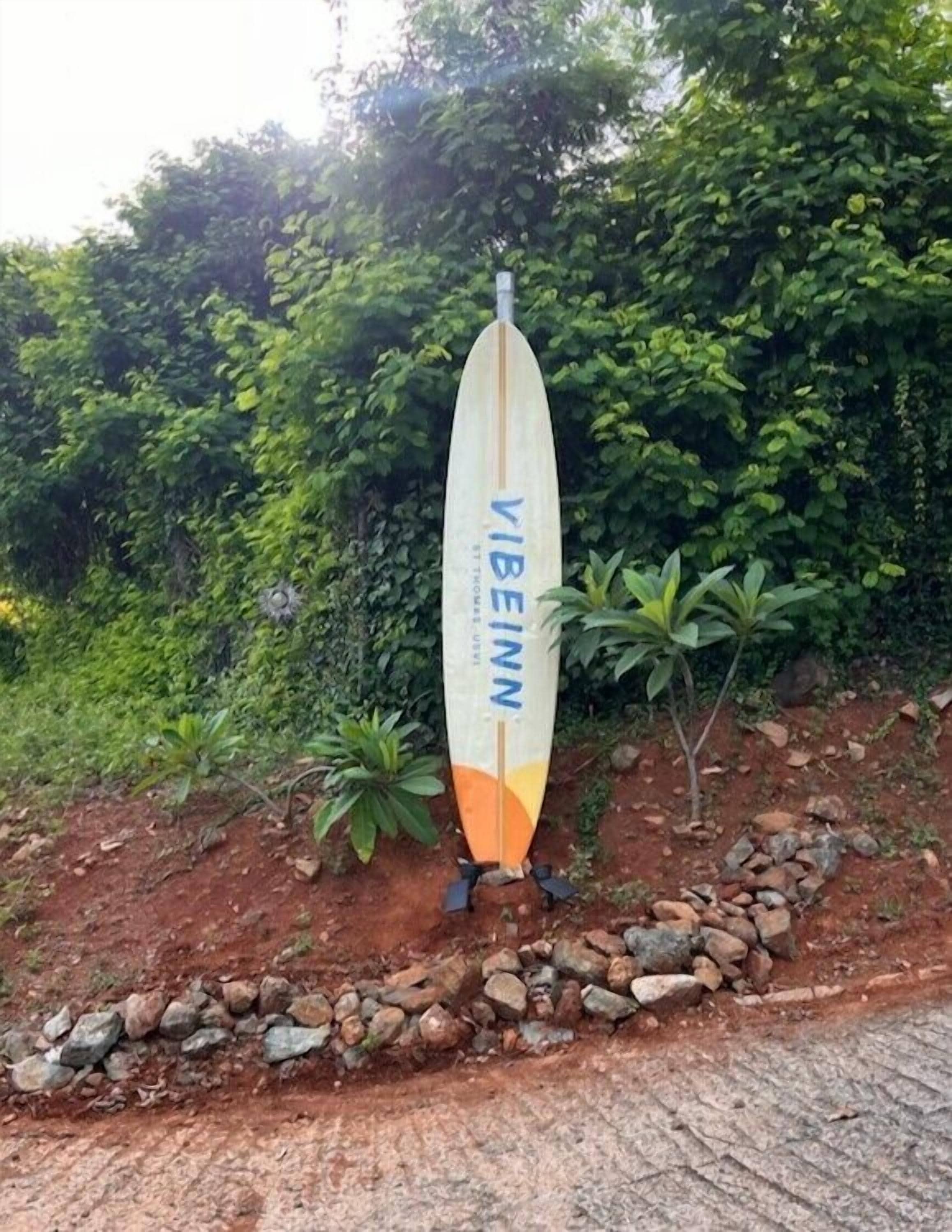 A surfboard with the text "VIBEINN" standing upright on a rocky surface with a backdrop of dense greenery.