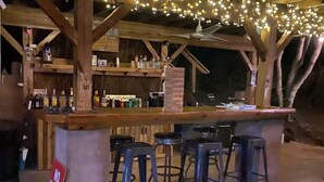 Ein Außenbereich mit Bar, der einen Beton-Tresen, Holzbalken und Lichterketten aufweist. Vier Barhocker stehen bereit, und ein Schild mit der Aufschrift 'NO DRINKS BEYOND THIS POINT'.