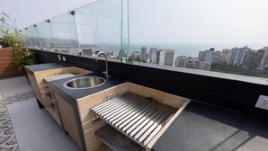 Une cuisine aménagée sur le toit, dotée d’un évier en acier inoxydable, d’un barbecue intégré et d’une véranda en verre offrant une vue panoramique sur la ville.