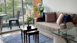 A modern living room with a glass dining table, a sofa with cushions, a coffee table, and a large window offering a city view. - Modern 1Bd Apt in the Best of Barranco Miraflores (Lima)
