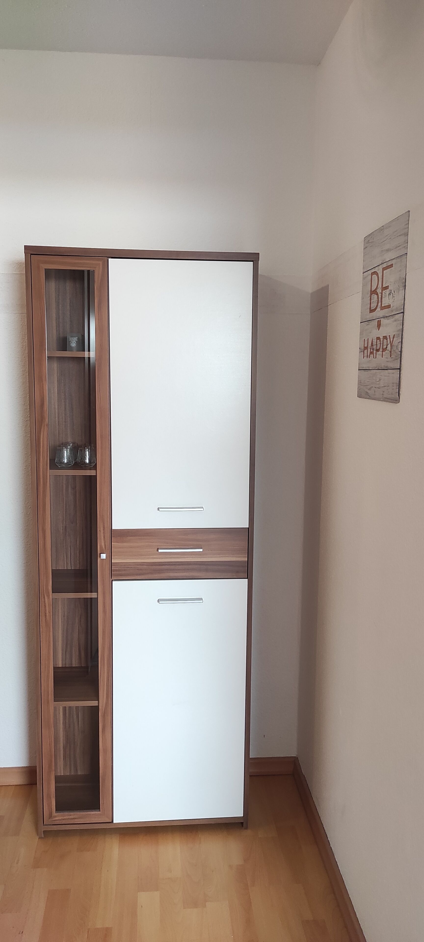 Ein Zimmer mit einem Holzschrank mit mehreren Einlegeböden und Schubladen, einer weißen Kommode und einem motivierenden Zitat an der Wand.