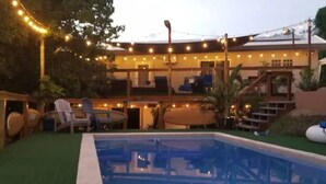 A pool area with a clear pool, surrounded by a deck with chairs and a table. There are string lights above the deck and a building in the background. - Surfer Shack 2 King Bedrooms (1 is a Loft) (East End)