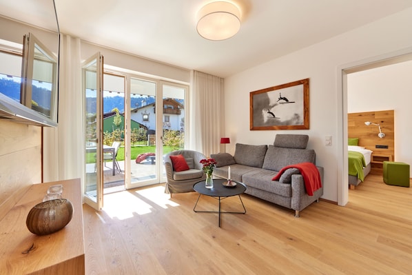 A modern living room with a sofa, armchair, and coffee table. There is a painting on the wall and a window with a view of houses and mountains.