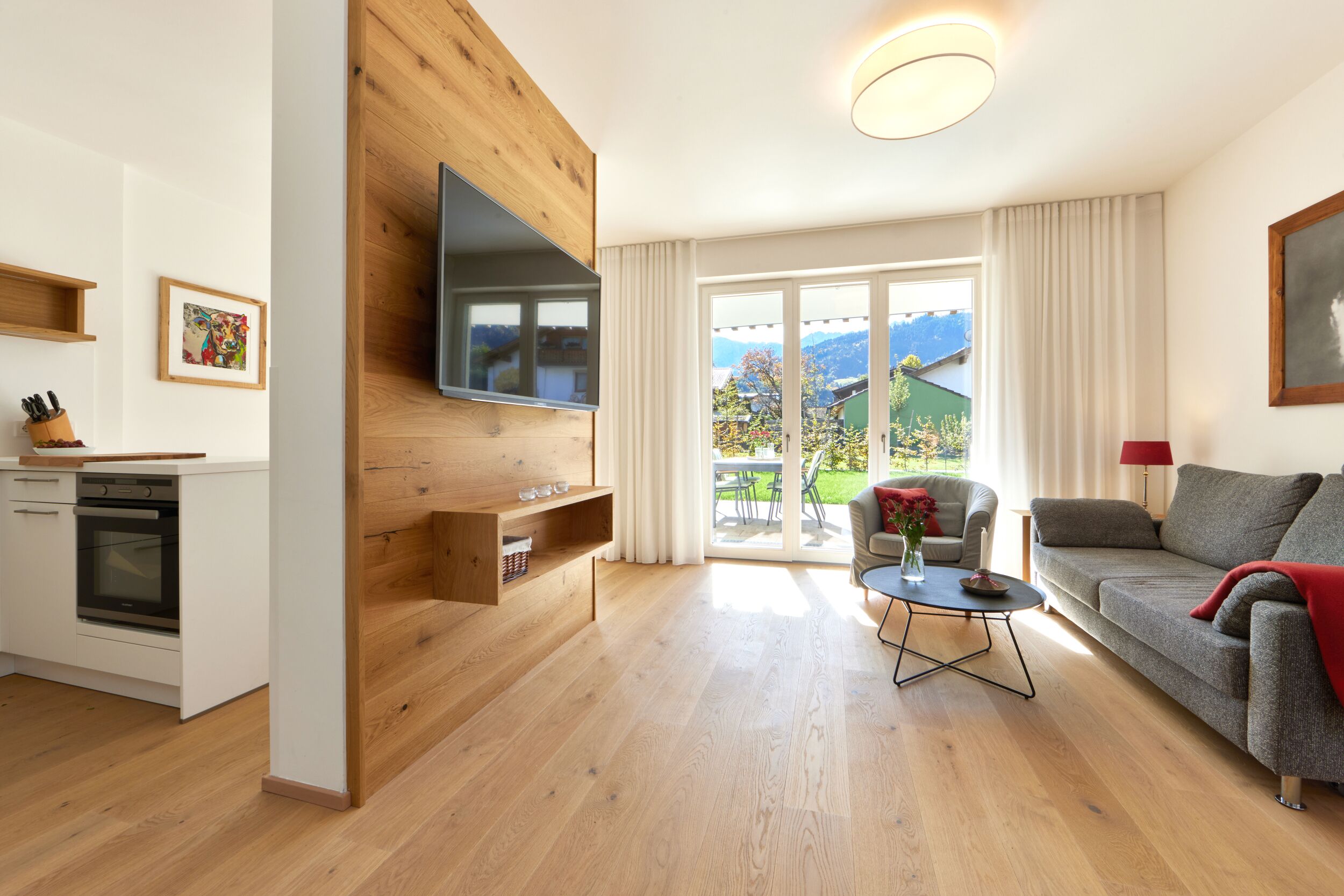 Ein modernes Wohnzimmer mit einer Holzwand, einem Flachbildfernseher, einer Couch, einem Couchtisch und einem Essbereich mit Bergblick.