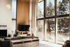 A modern living room with a large TV, a fireplace, and a view of trees through large windows.
