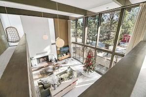 Un intérieur moderne avec une cheminée, un coin salon et de grandes fenêtres donnant sur des arbres et un bâtiment rouge.