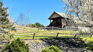 A wooden cabin with a sloped roof, surrounded by a wooden fence, set in a grassy area with trees and a clear sky.