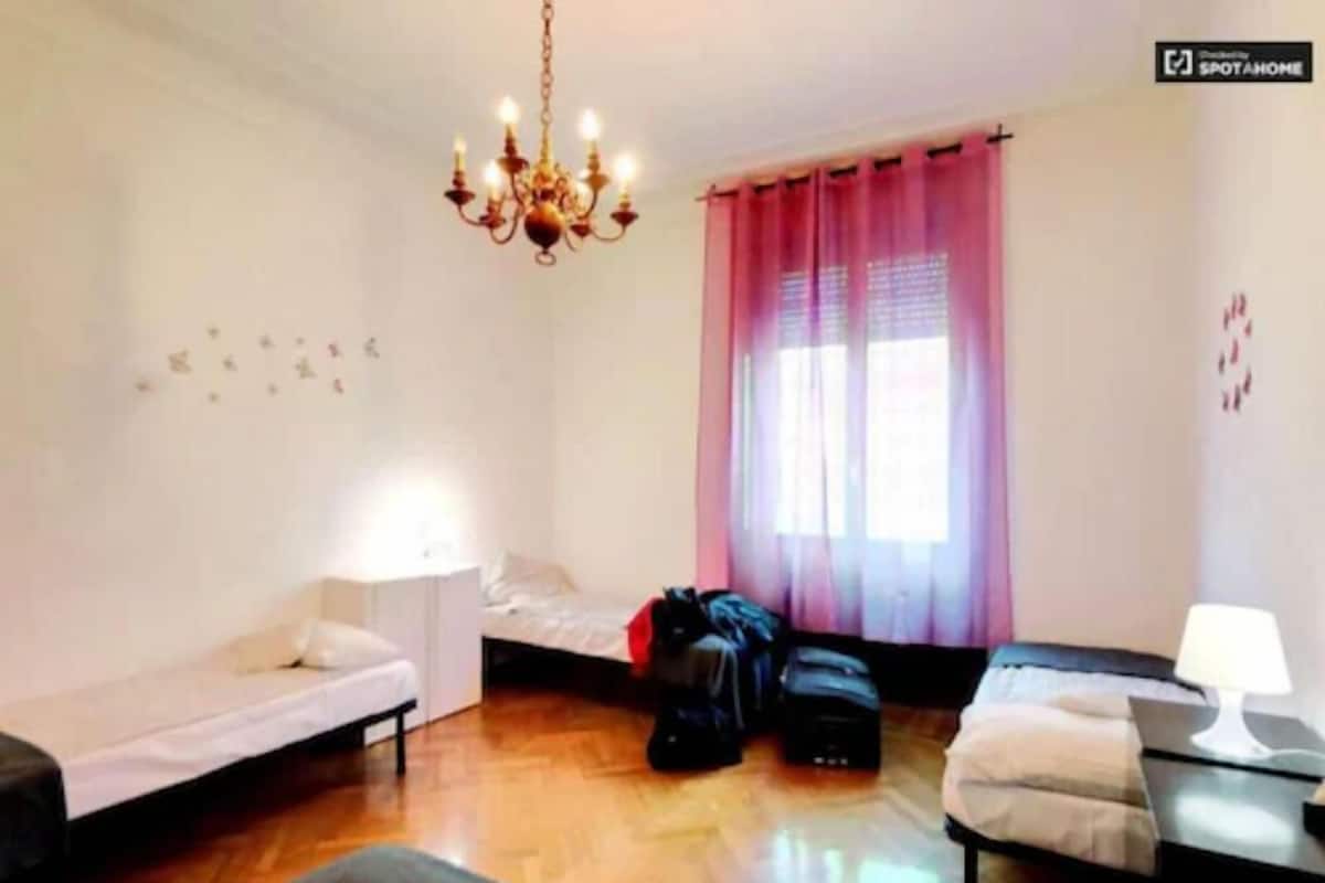 A small, well-lit room with a bed, a desk, a chair, a suitcase, and a chandelier.
