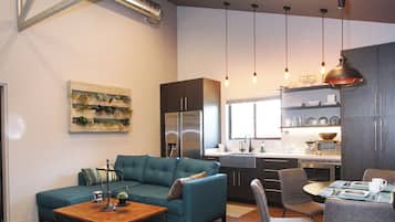 A modern living room with a sofa, coffee table, and dining area with chairs. There is a kitchen in the background with a sink and appliances.