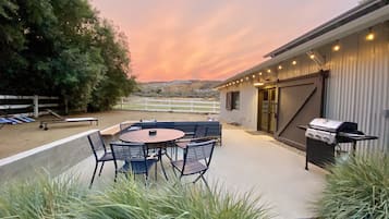 An outdoor patio with a round table and chairs, a barbecue grill, and a view of a building with a white fence in the background.