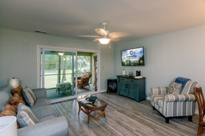 A living room with a sofa, armchair, and a coffee table. There is a television mounted on the wall and a ceiling fan.