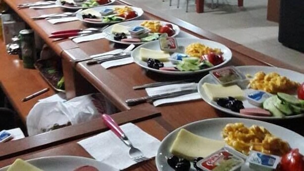 A buffet setup with various dishes and utensils on a long table, surrounded by chairs and tables in a dining area.