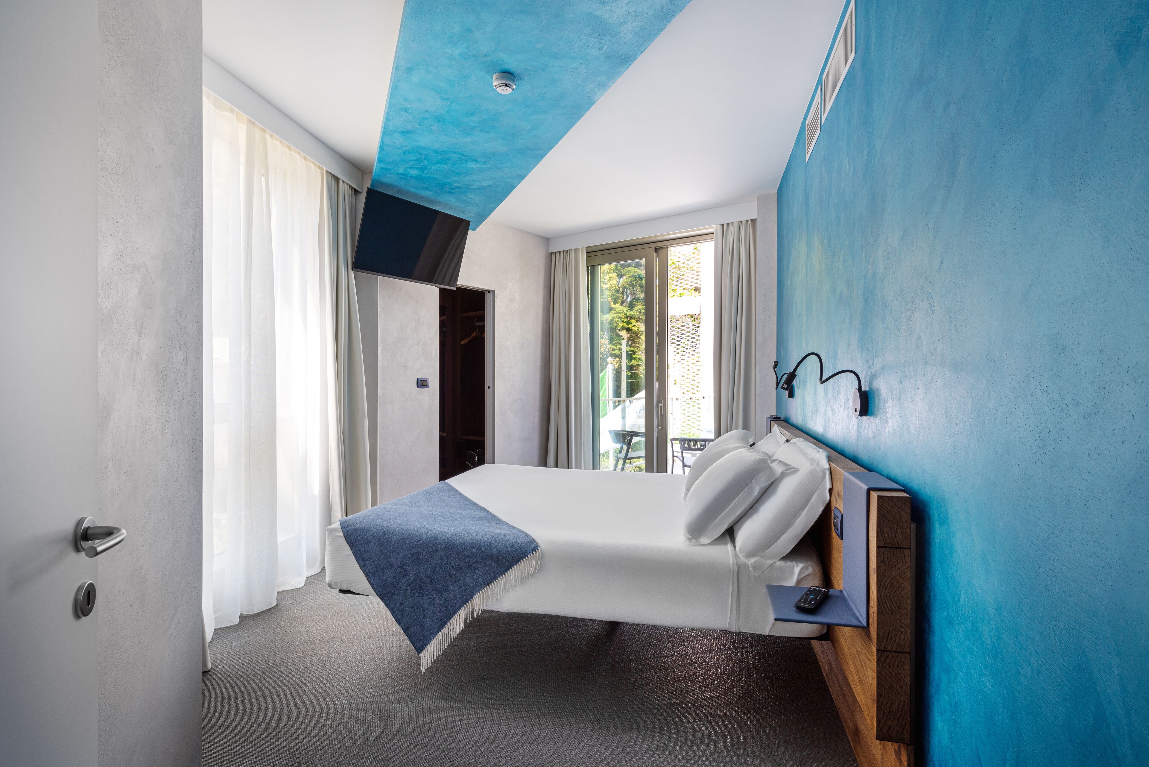 Une chambre d’hôtel moderne avec un lit, une table de chevet, un plafond bleu et une vue sur l’extérieur.