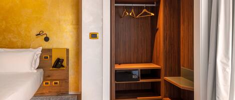 A hotel room with a bed, a bedside table with a phone, a small shelf, a wooden closet with hangers, and a grey carpet.
