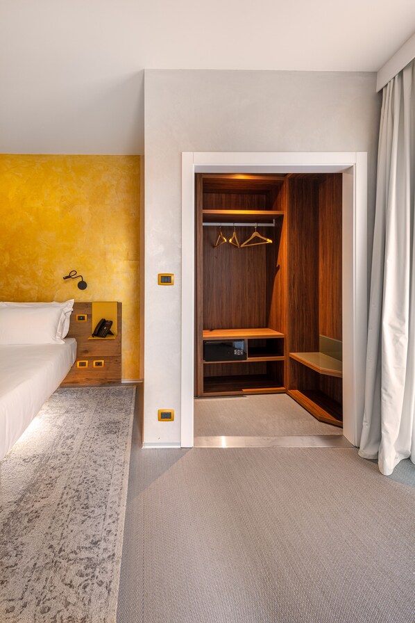 A hotel room with a bed, a bedside table with a phone, a small shelf, a wooden closet with hangers, and a grey carpet.