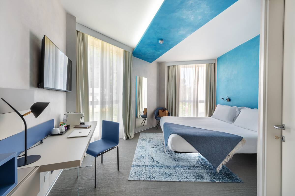 A modern hotel room with a bed, desk, chair, and a large window with curtains.
