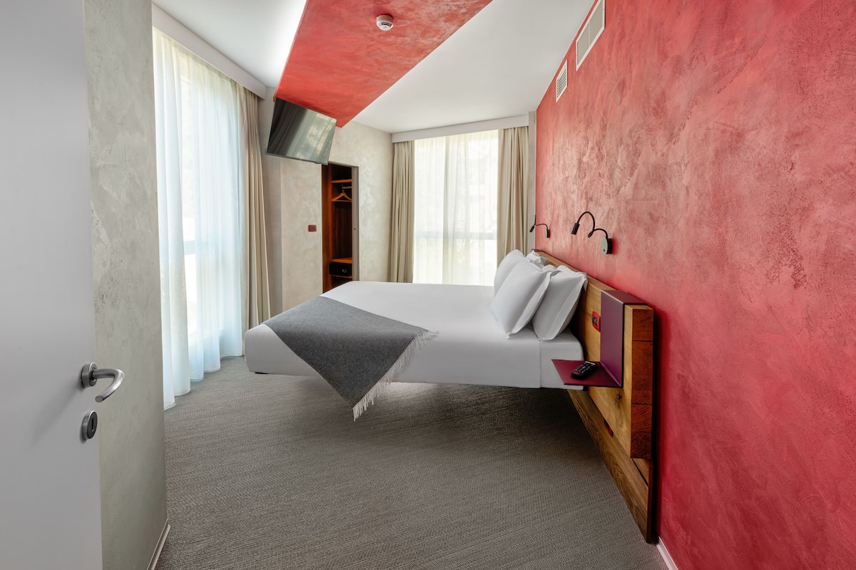 A modern hotel room with a bed, a wooden headboard, a wardrobe, and a large window with curtains.