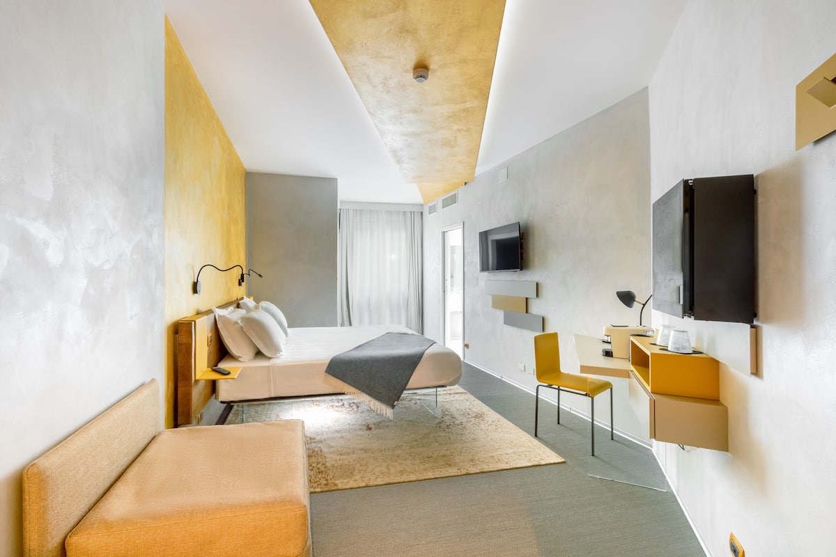 A modern hotel room with a bed, a desk, a chair, a TV, and a large window.