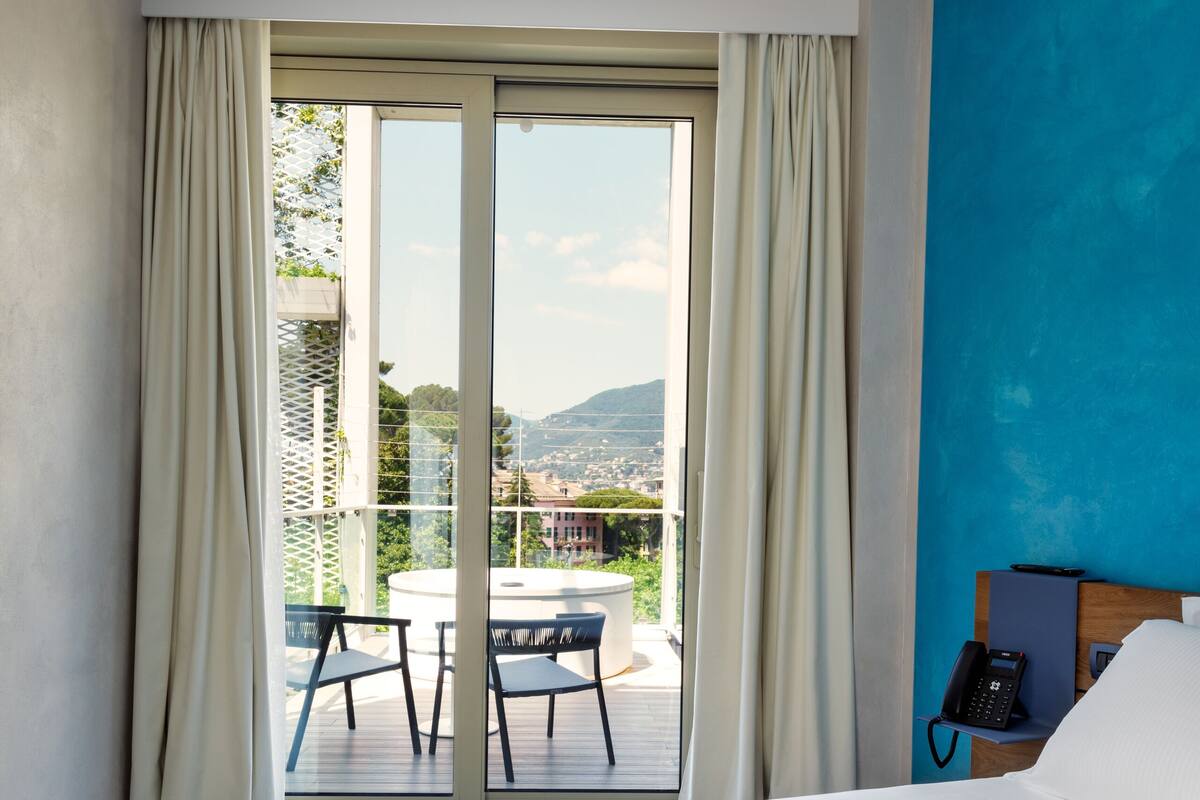 A hotel room with a large bed, a balcony with a table and chairs, and a view of the city and mountains.