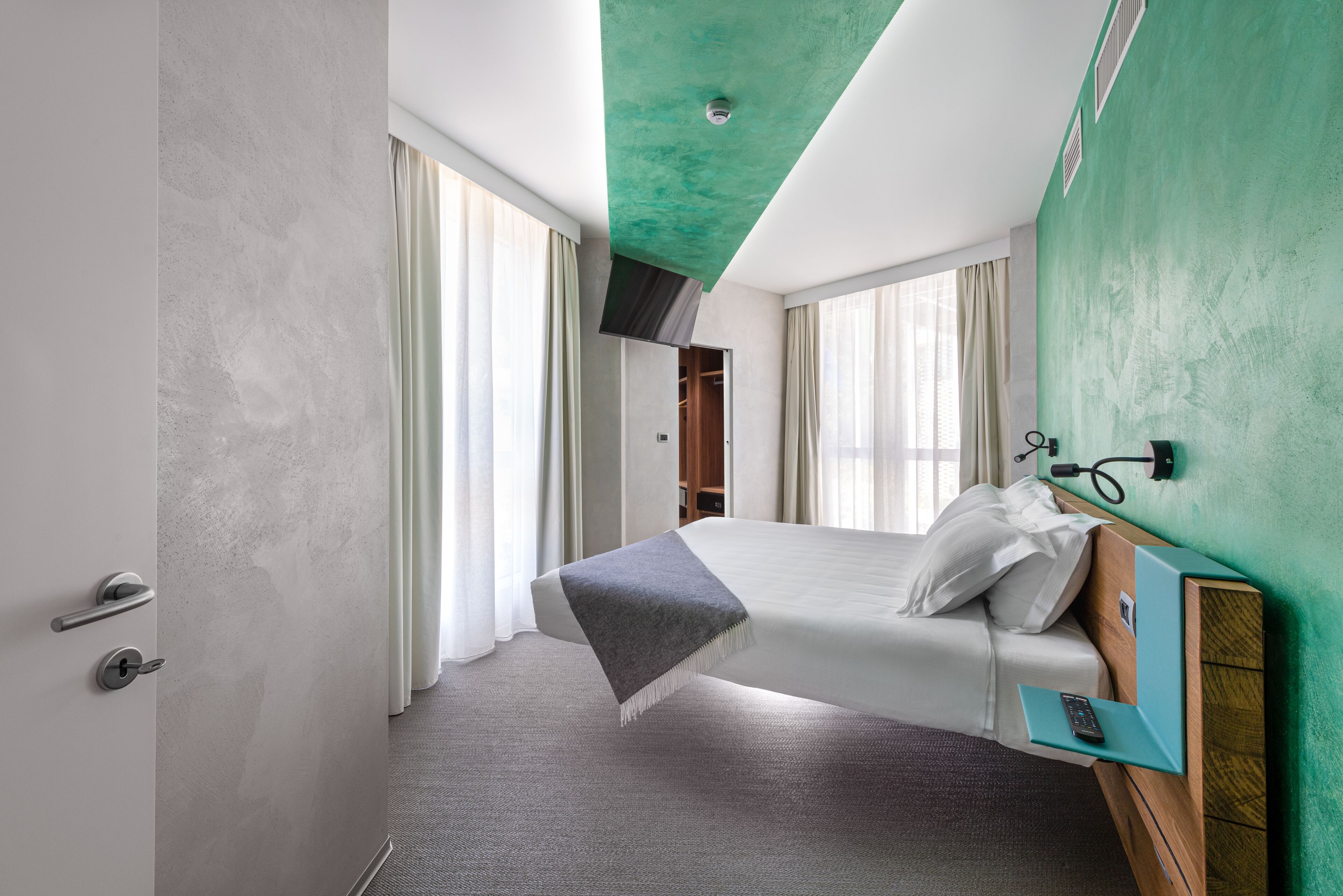Una habitación de hotel moderna con una cama grande, un cabecero de madera, una mesita de noche con un control remoto y una pared de acento verde.
