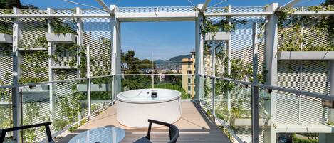 A balcony with a glass table and chairs, a hot tub, and a view of the city.