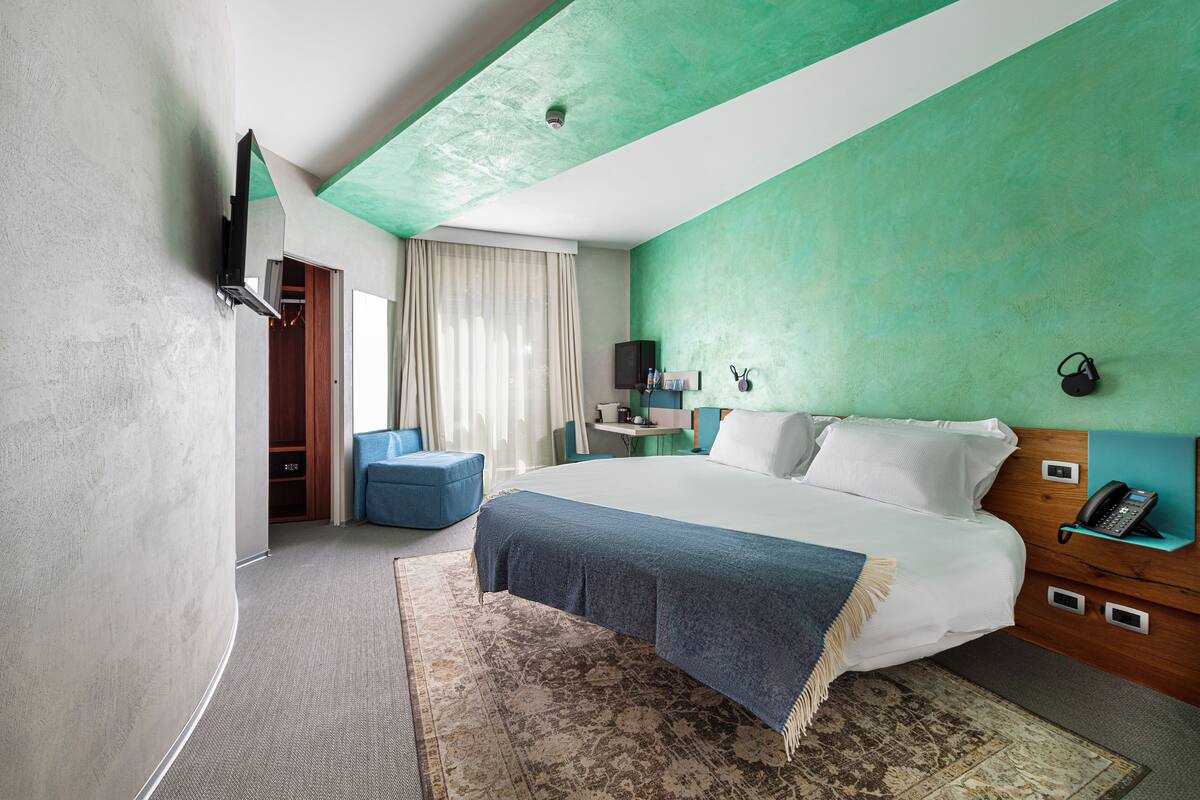 A modern hotel room with a large bed, a desk, a chair, and a wardrobe.