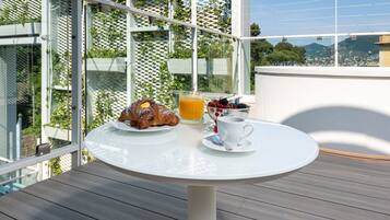 Ein runder Tisch mit Frühstücksangeboten, darunter Croissants, ein Glas Orangensaft und eine Tasse Kaffee, auf einem Balkon mit Blick auf Grünflächen und Gebäude.