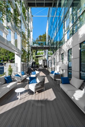 Un área de estar al aire libre con sofás blancos y cojines azules, una terraza de madera y un frente de cristal del edificio.