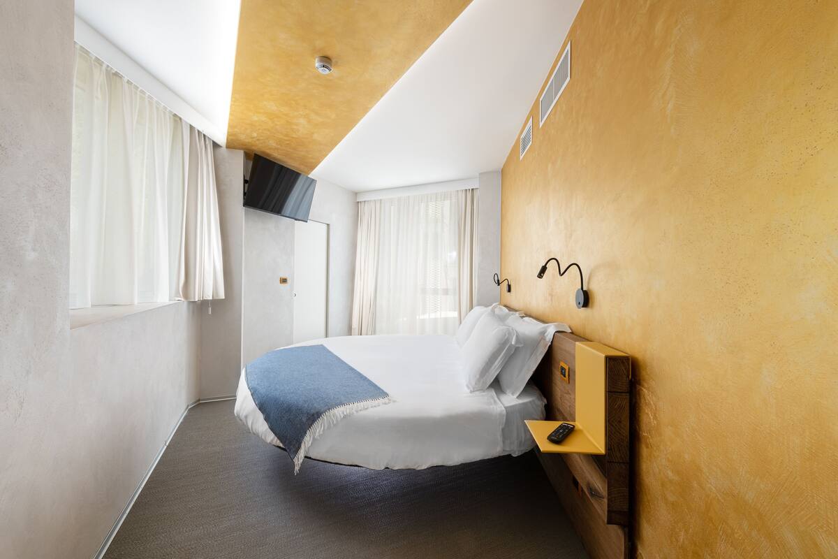A hotel room with a bed, a bedside table, a wall-mounted lamp, and a window with curtains.