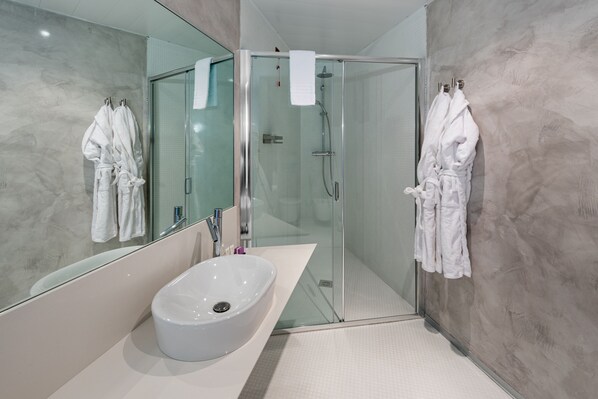 Un baño moderno con ducha envuelta en vidrio, un lavabo blanco y dos albornoces blancos colgados en ganchos.