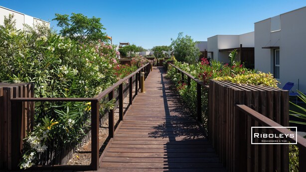 Ein hölzerner Weg schlängelt sich durch einen Gartenbereich mit verschiedenen Pflanzen und Blumen, gesäumt von modernen Gebäuden mit weißen Wänden und Balkonen.
