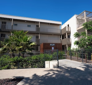 Un edificio moderno con varios balcones, entrada protegida y abundante vegetación.