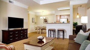 Sala de estar con televisor, mesa de centro y sofá. Se observa un comedor con mesa y sillas. Se ve una cocina al fondo.