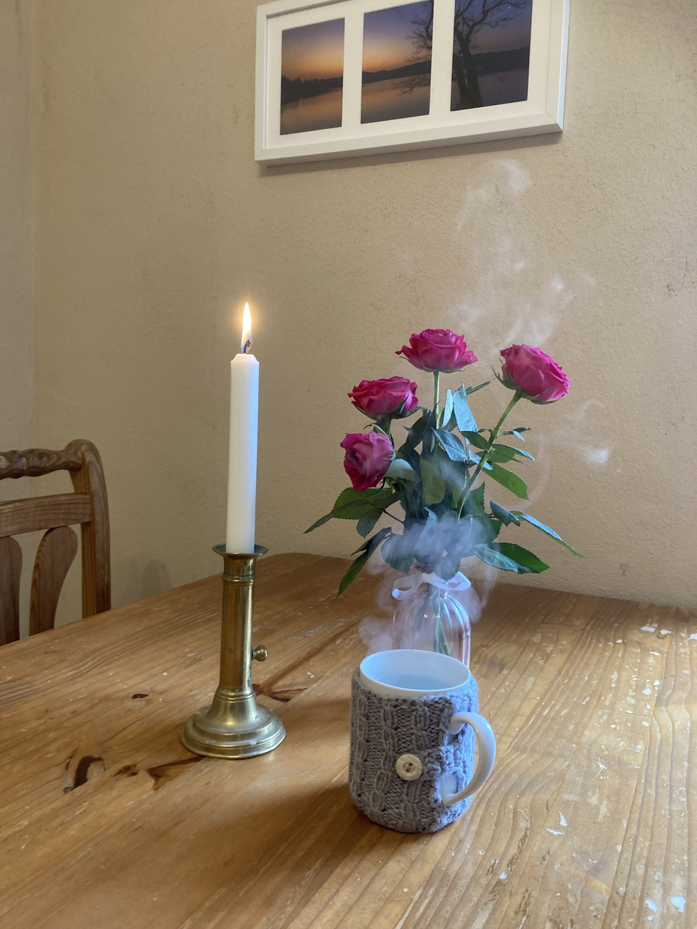 A table with a lit candle, a vase of pink roses, and a ceramic mug.