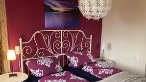 Une chambre avec un lit en métal blanc, une literie à motifs violets et blancs, une table ronde avec une lampe et un luminaire suspendu.