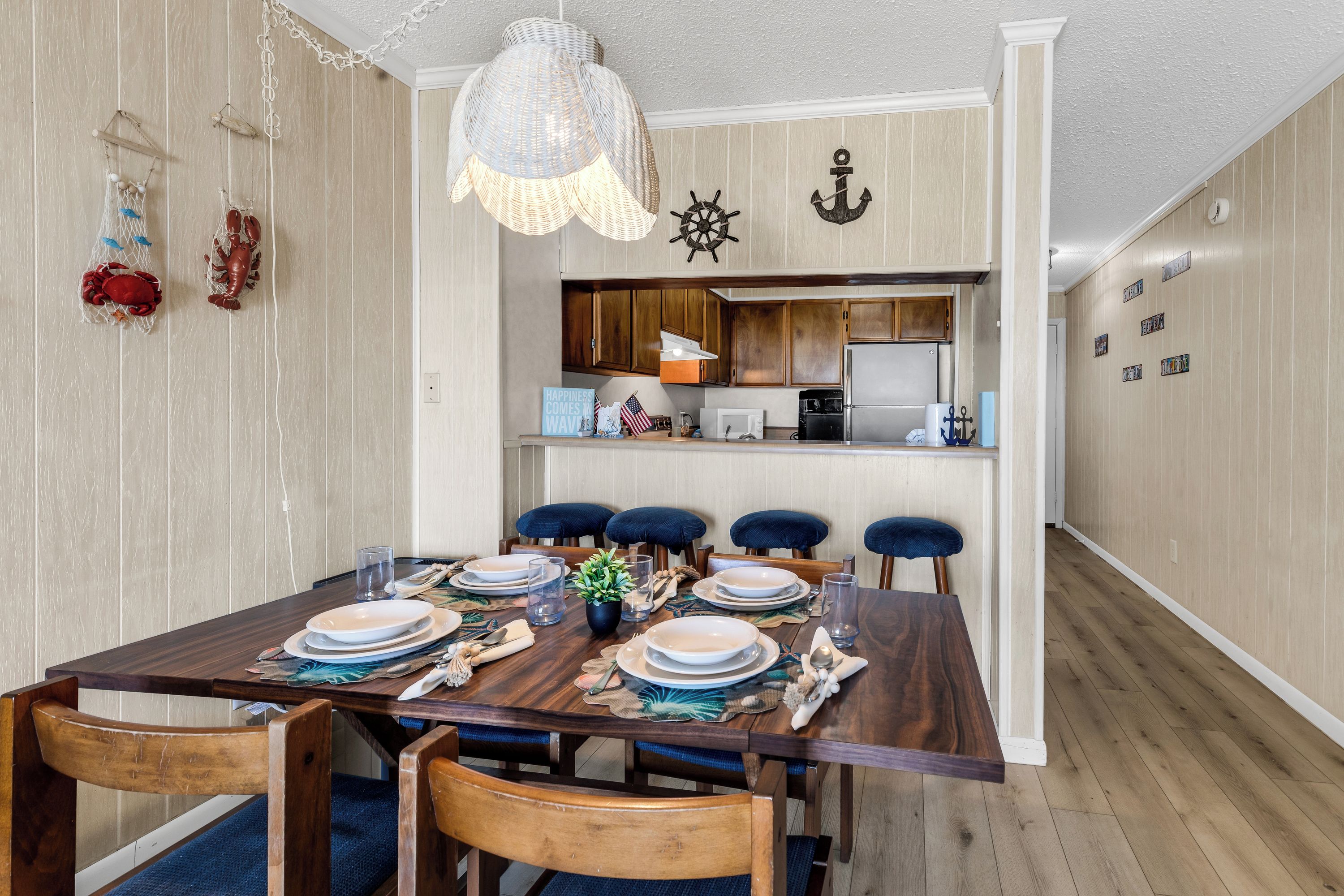 A dining area with a wooden table set for six, a kitchen in the background, and nautical-themed wall decorations.
