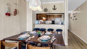 A dining area with a wooden table set for six, a kitchen in the background, and nautical-themed wall decorations.