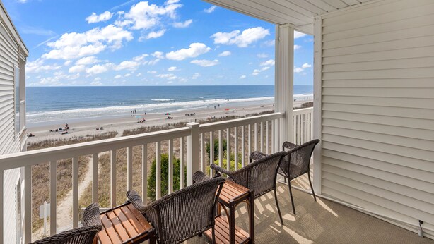 Ein Balkon mit Korbstühlen und einem Tisch mit Blick auf einen Strand, auf dem sich Personen und Sonnenschirme befinden.
