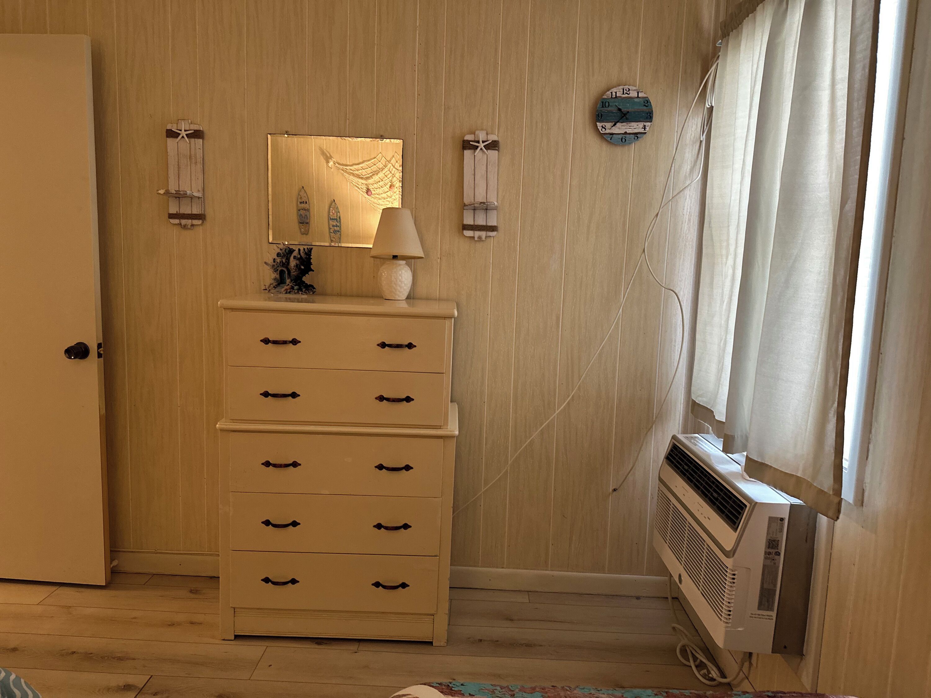 Una habitación con un armario de madera, una lámpara de pared, un reloj y una ventana con cortinas.