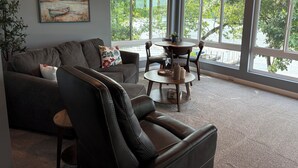 A living room with a couch, a leather chair, a round table, and large windows offering a view of trees and a body of water. - PEACEFUL HOME-4 BEDROOMS, 3 KING BEDS, 4 BATHROOMS, EASY PARKING & A BOAT SLIP! (Camdenton)