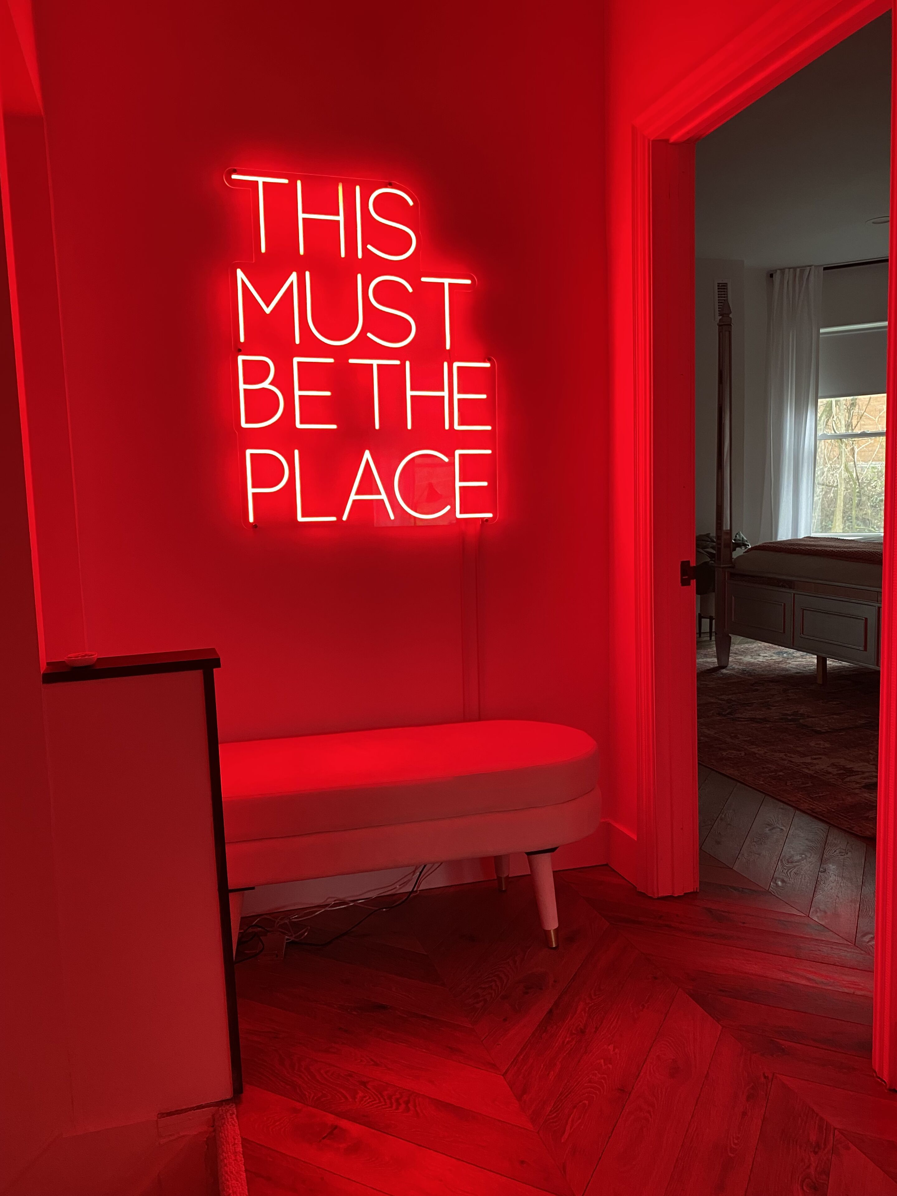 Ein Zimmer mit einer Neonreklame, auf der "THIS MUST BE THE PLACE" steht, einer Bank und Blick auf ein Schlafzimmer durch eine offene Tür.