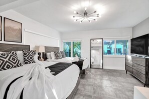 Una habitación de hotel moderna con una cama grande, televisión, cómoda y un baño visible a través de una puerta de cristal.