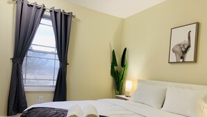 Habitación de hotel con una cama, una mesita de noche con lámpara, una ventana con cortinas, una planta y un cuadro en la pared.