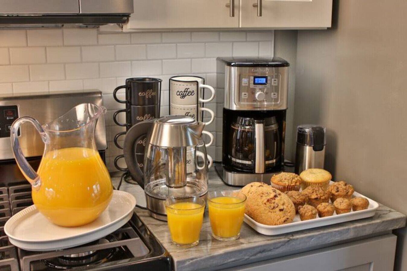 A kitchen with a coffee maker, a coffee pot, a pitcher of orange juice, and a tray of muffins.