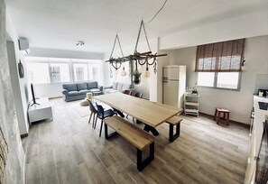 Tilava olohuone, jossa on puinen ruokapöytä, penkit ja sohva. Huoneessa on myös amppelikasveja ja kattovalaisin.