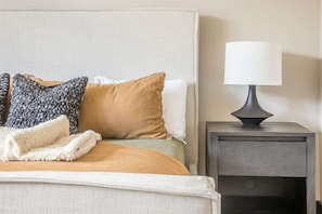 Una cama bien hecha con edredón beige y blanco, un plaid gris tejido y una manta blanca. Se observa una mesita de noche con una lámpara blanca y un tocador de color oscuro.